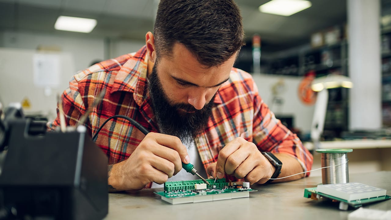 Bearded technician soldering electronic components on a circuit board at a workbench.
