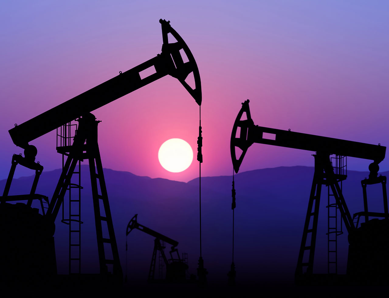 Silhouettes of oil pump jacks operating at sunset with mountains in the background.